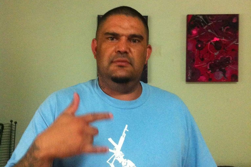 A large-statured man with short hair, wearing a blue t-shirt, makes a 'shakkas'-style hand gesture at the camera.