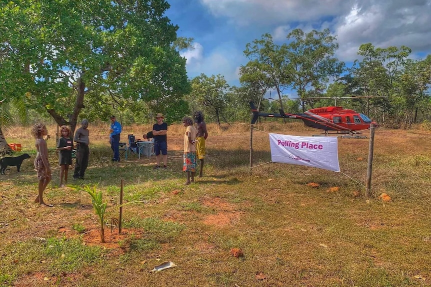 people gather at a remote polling station with a helicopter in the background. 