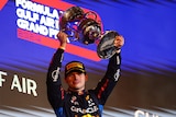 Max Verstappen, in his racing suit, lifts a gold trophy over his head in celebration.