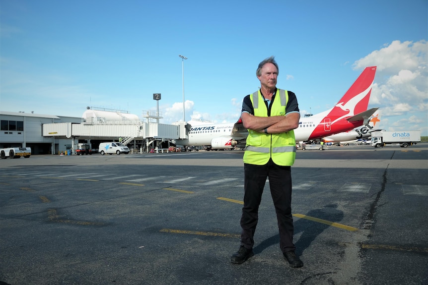 Man in hi-viz vest standing arms crossed on the apron at an airport with a qantas plane behind.