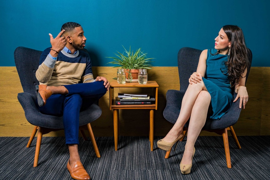 A man and woman sitting down talking, the man has he hand in the air, the woman looks unimpressed