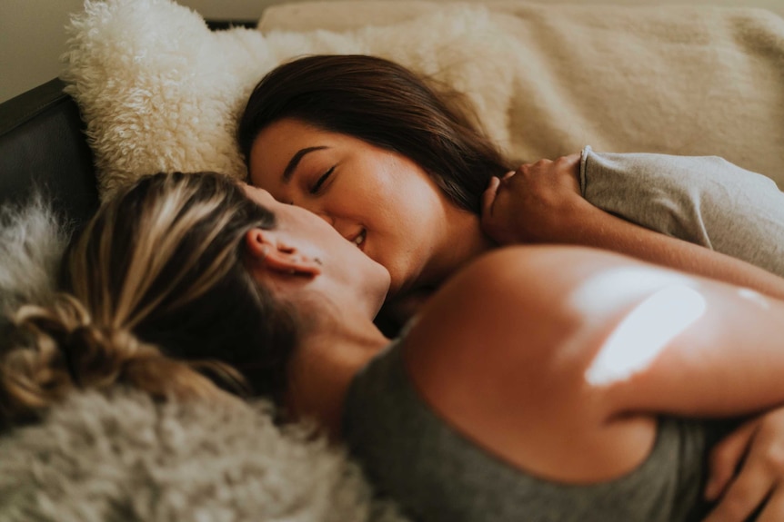 Two women in bed lying down and holding each other together and kissing.