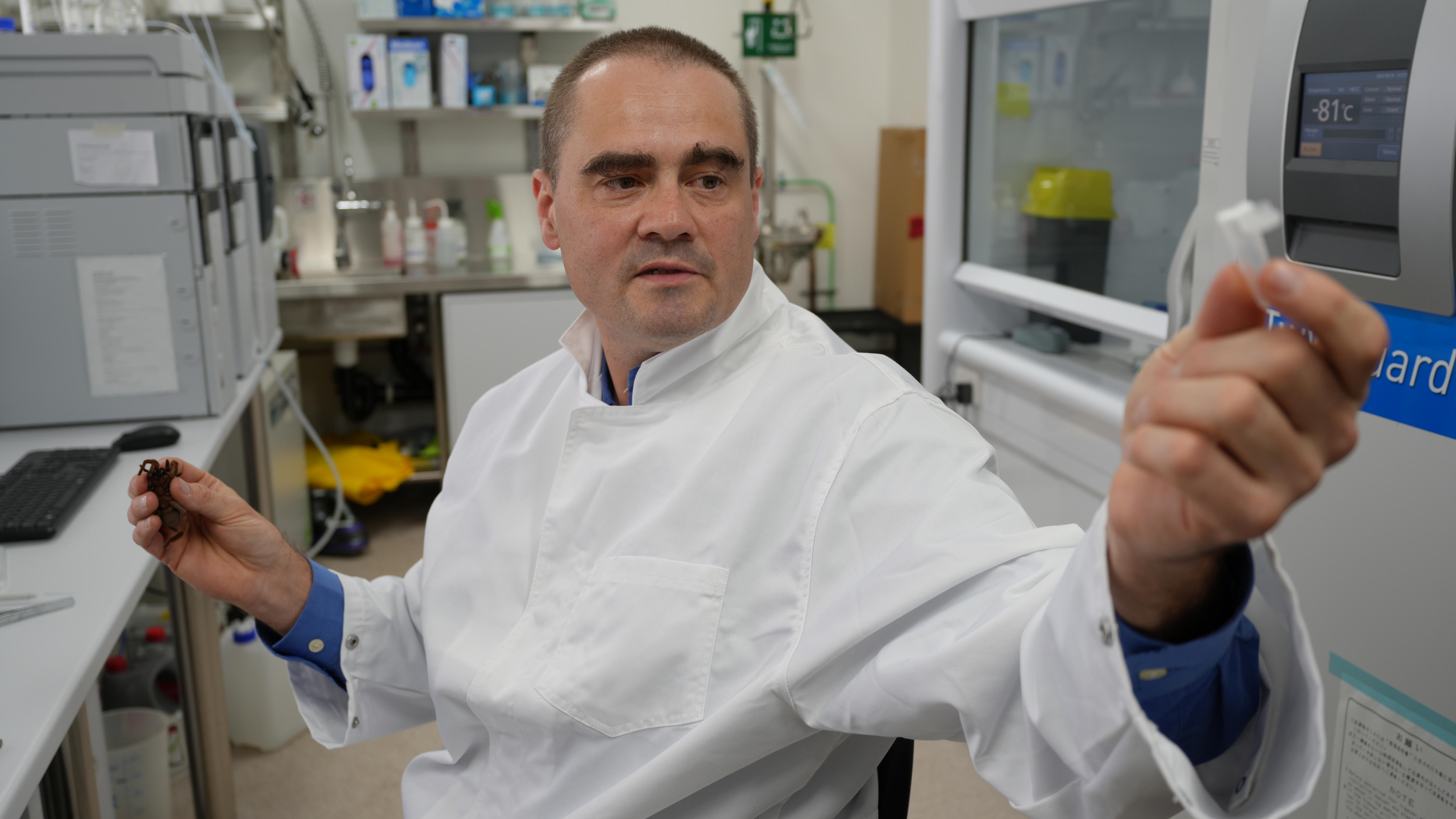 A dark-haired man in a lab coat holds up a sample container.
