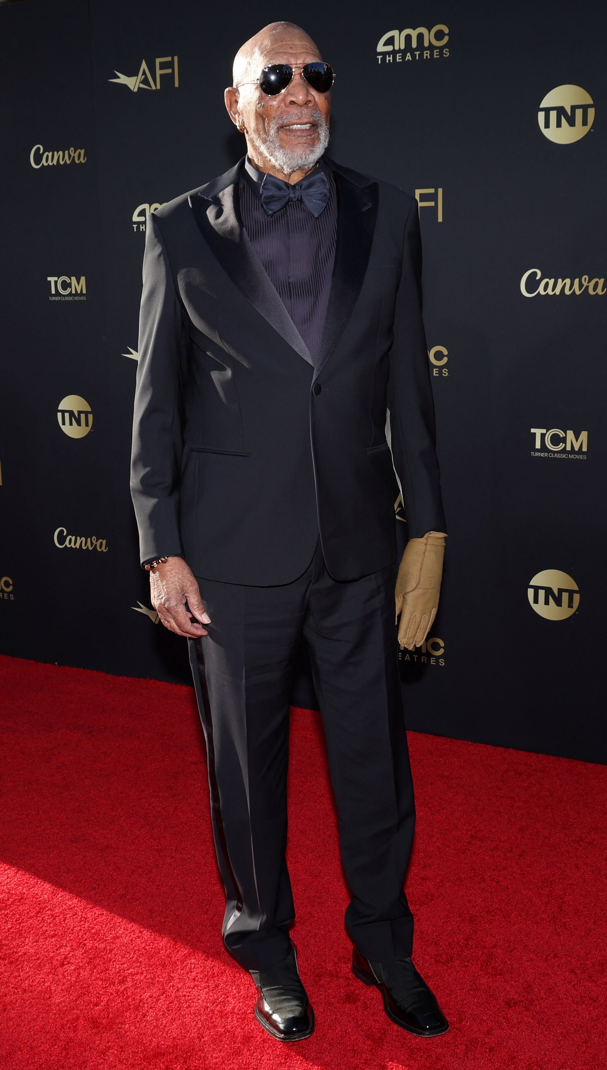 Morgan Freeman wearing a black suit with black aviator sunglasses and a black shirt and bow tie and one brown leather glove