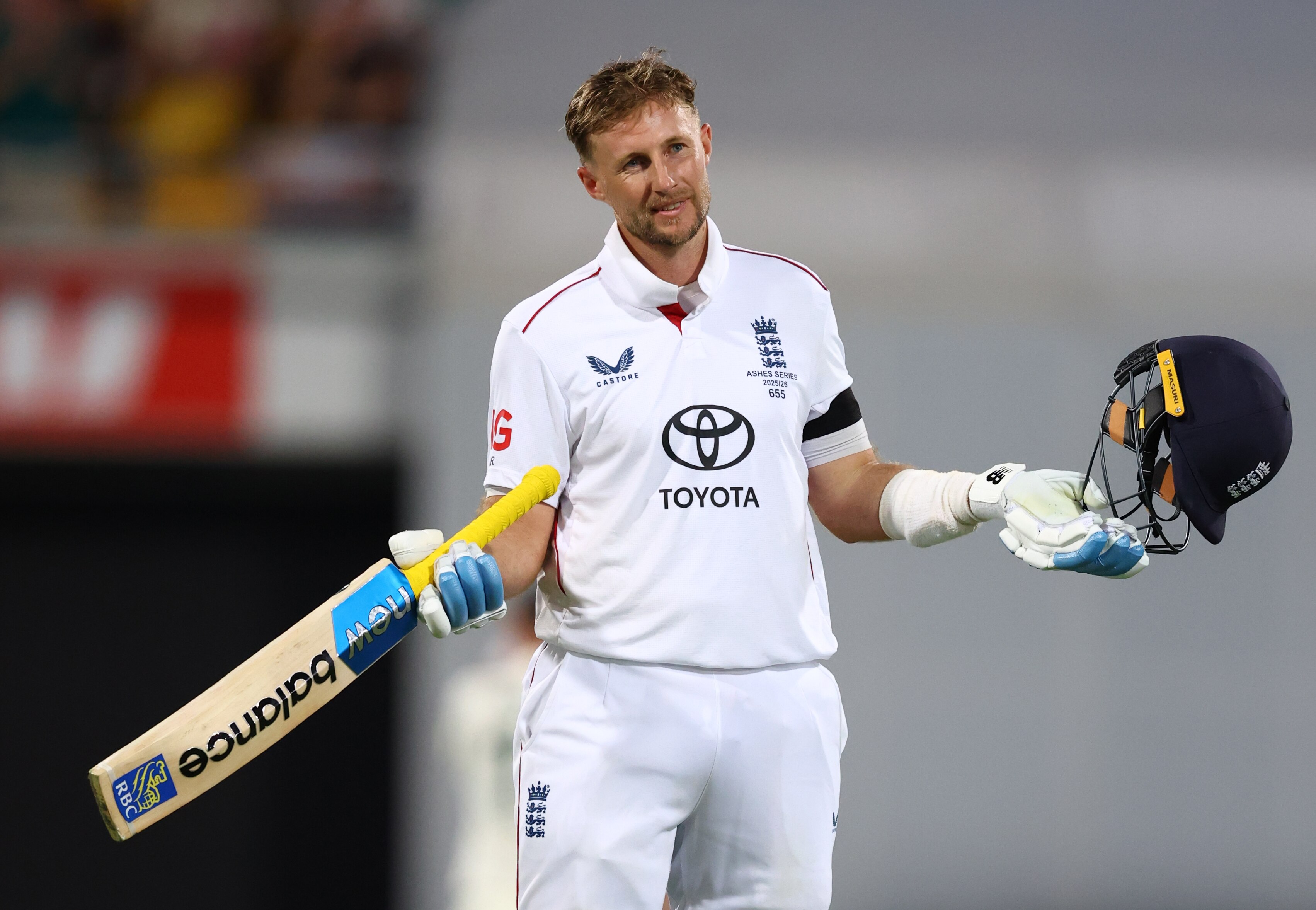 Un jugador de críquet vestido de blanco sostiene su casco y su bate, sonriendo