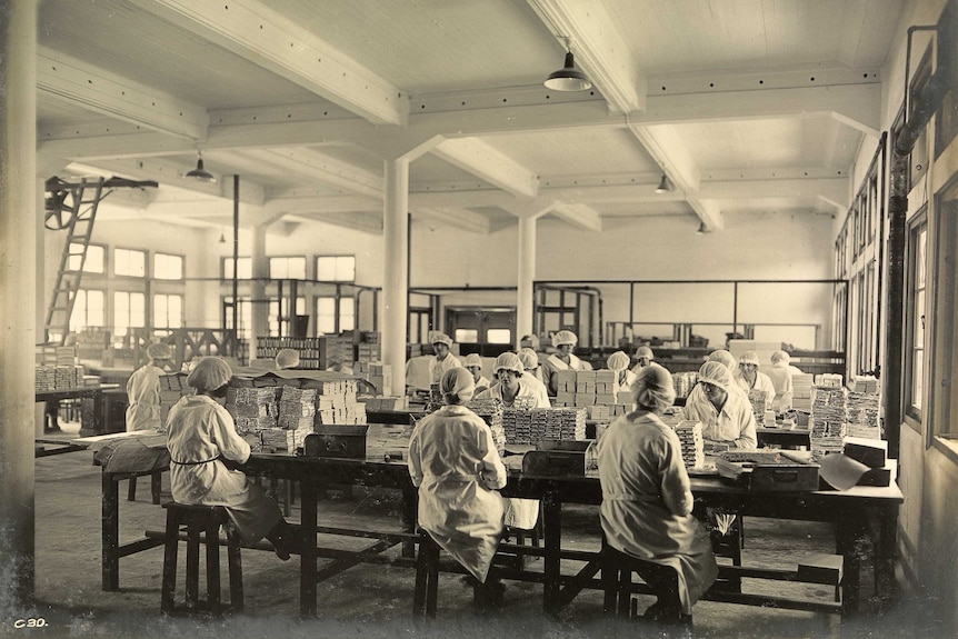 Sepia photo of a factory floor with women in white coats and hairnets wrapping chocolate.