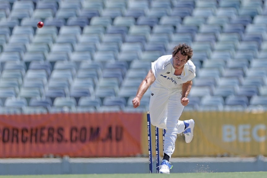 A young men's fast bowler is pictured in his follow-through from a delivery as the ball flies down the pitch.  