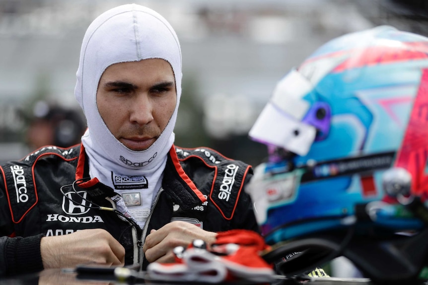 A man wearing an open-faced balaclava zips up his race suit with his helmet in the foreground