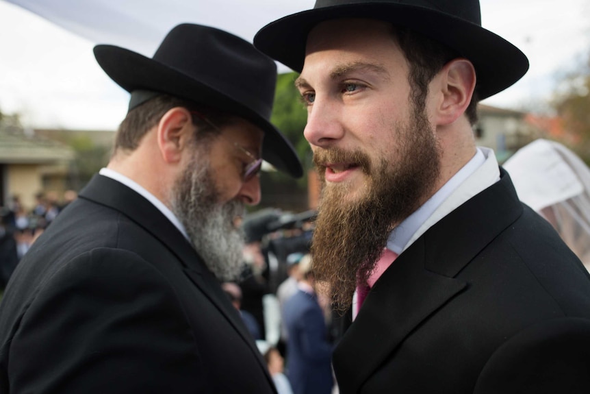 A young Jewish man, flanked by an older one, each in identical beard and black hat.