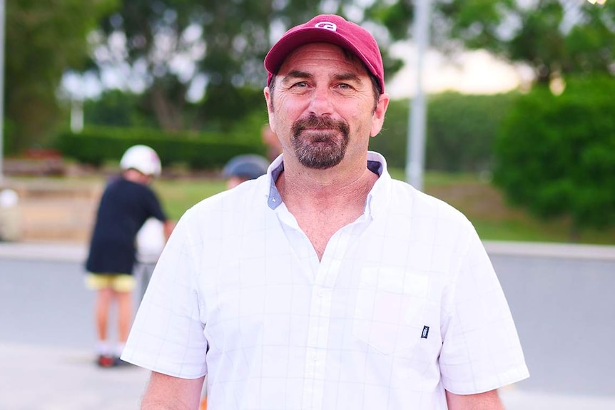 Man with white shirt and red cap standing in front of blurred skate park.