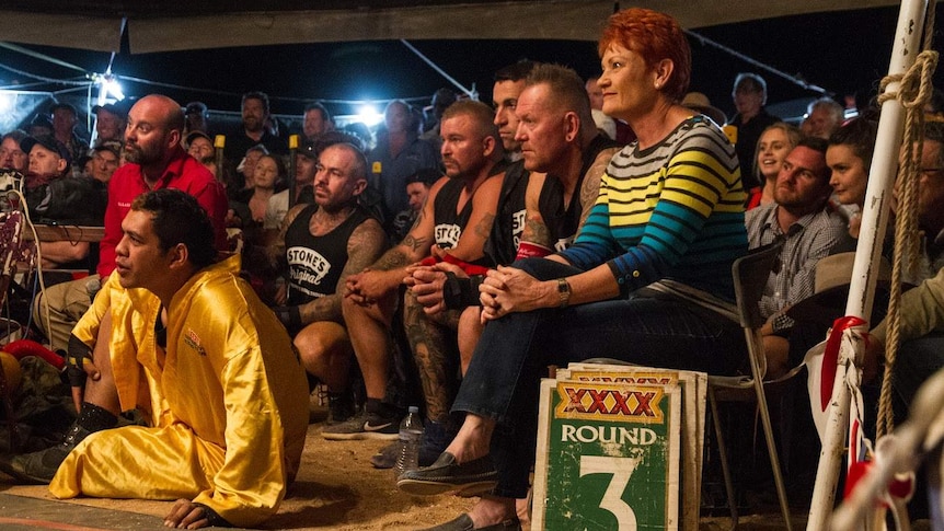 Pauline Hanson sits next to tattooed men in a crowd all watching something out of sight. It's nighttime and a sign says Round 3.