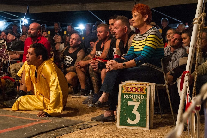 Pauline Hanson sits next to tattooed men in a crowd all watching something out of sight. It's nighttime and a sign says Round 3.