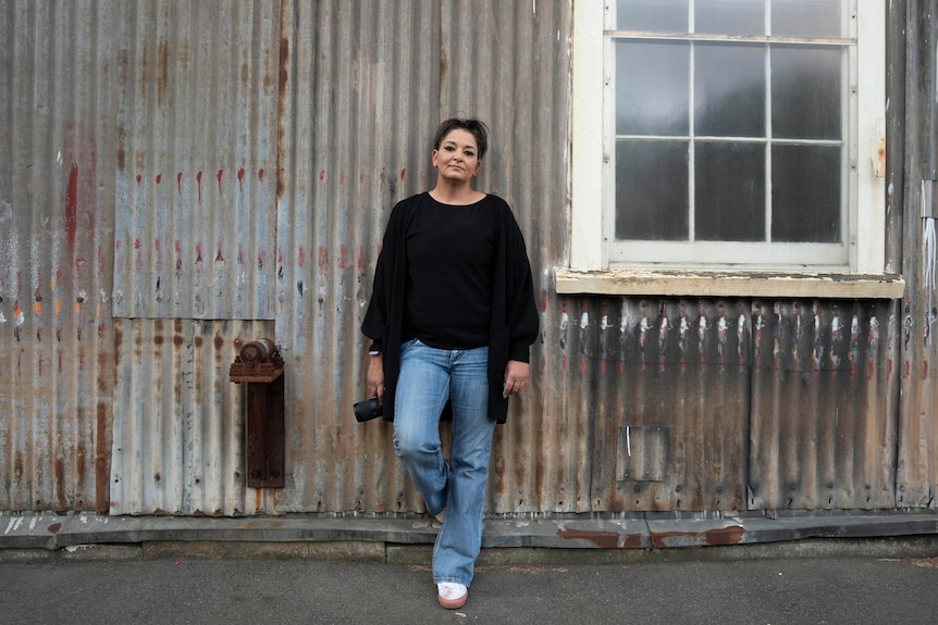 A woman wearing a black top and blue jeans leans against a rusty corrugated iron wall, holding a camera