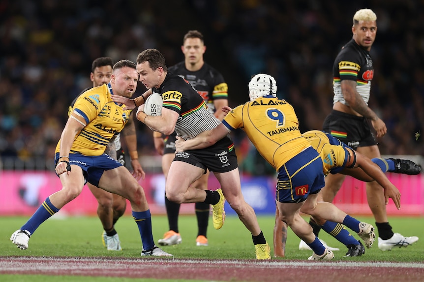 Un jugador de Penrith Panthers NRL sostiene el balón con una mano y lo esquiva con la otra mientras intenta romper dos tacleadas.