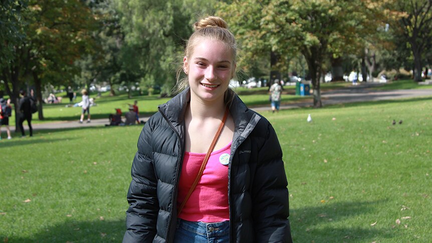 A 13-year-old girl in a black puffer jacket stands in a sunny park.