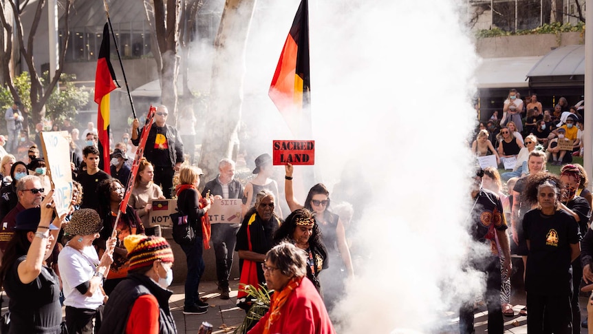 A group of people holding signs reading 'sacred land' and Aboriginal flags surround a smoking fire in a CBD park.