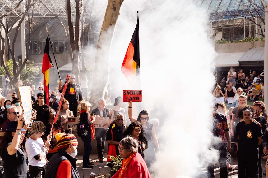 A group of people holding signs reading 'sacred land' and Aboriginal flags surround a smoking fire in a CBD park.