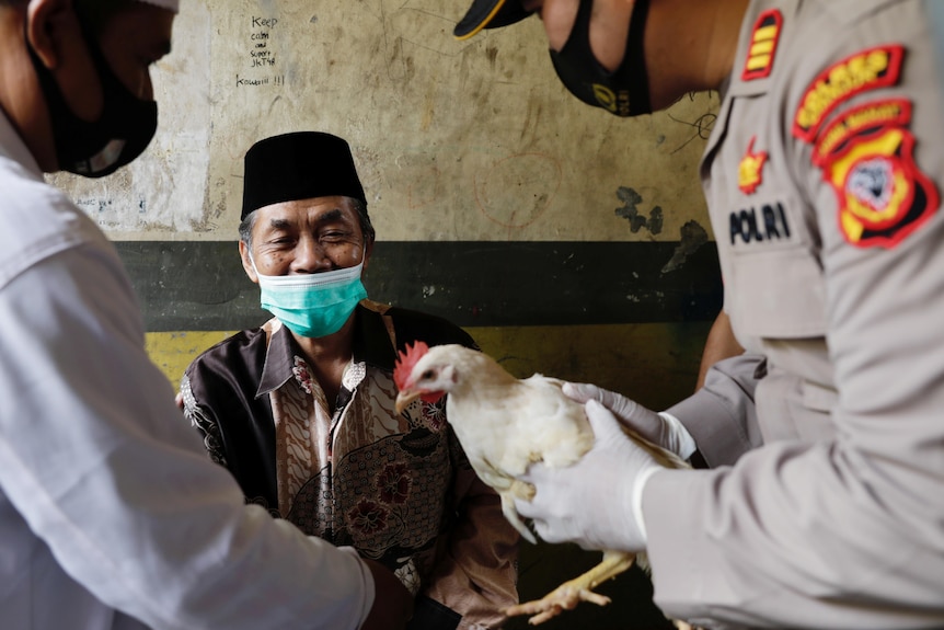 An Indonesian man in a face mask grins as two men hand him a chicken