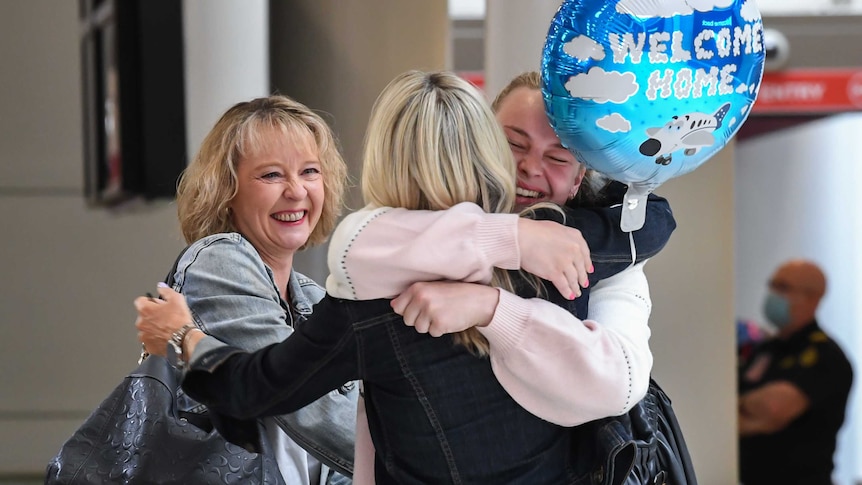 Two women hug one another as they meet in an airport arrivals area. One holds a balloon that says 'Welcome Home'.