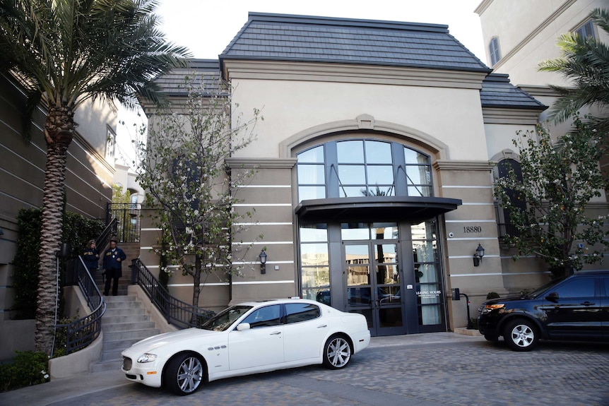 A brown apartment complex is seen with a white and black car parked out front. Two police officers stand to left on staircase.