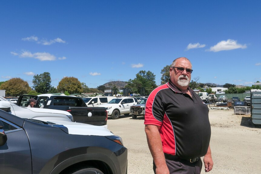 A serious middle aged man with paunch stands in front of cars, wears sunglasses, black and red t-shirt.