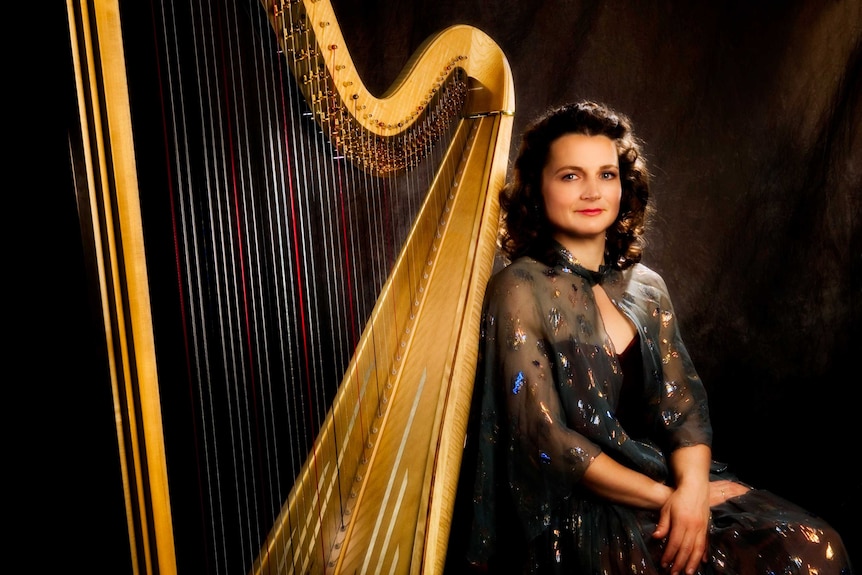 A woman in an evening gown sits next to a harp in a studio-type setting