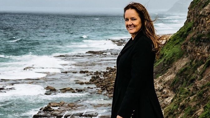 A woman with long hair, wearing a black coat, standing near the ocean.