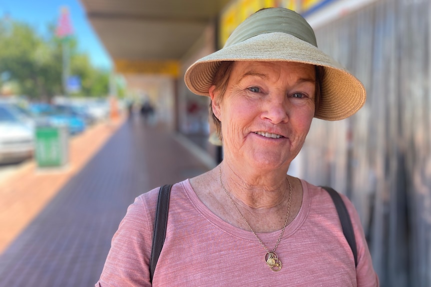 Smiling woman wearing pink shirt and beige visor, standing outside bank on footpath