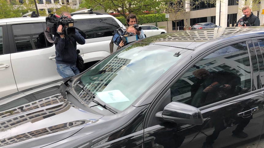 Unmarked Australian Federal Police car drives away from Home Affairs office surrounded by media