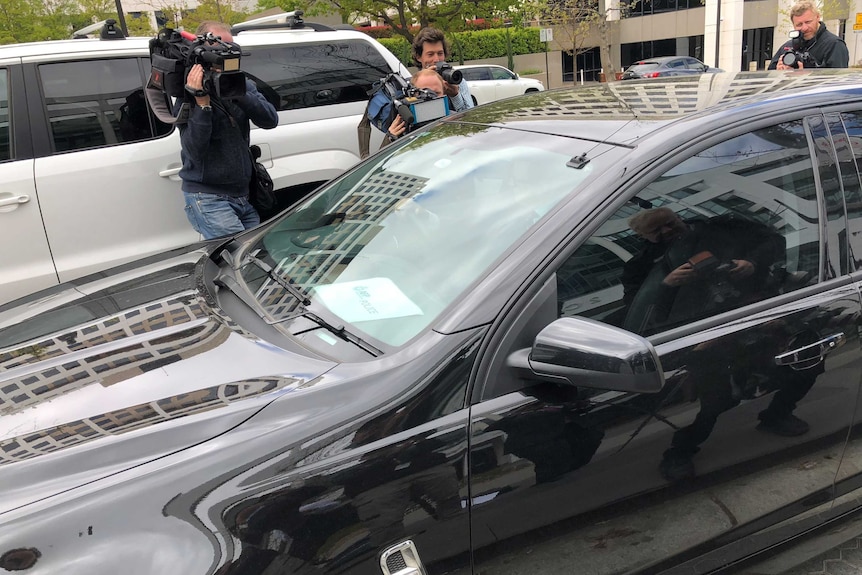 Unmarked Australian Federal Police car drives away from Home Affairs office surrounded by media