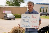 A middle-aged man stands next to a camping chair in a parking lot holding a sign: 'Goodbye Roe Thank God and Donald Trump'