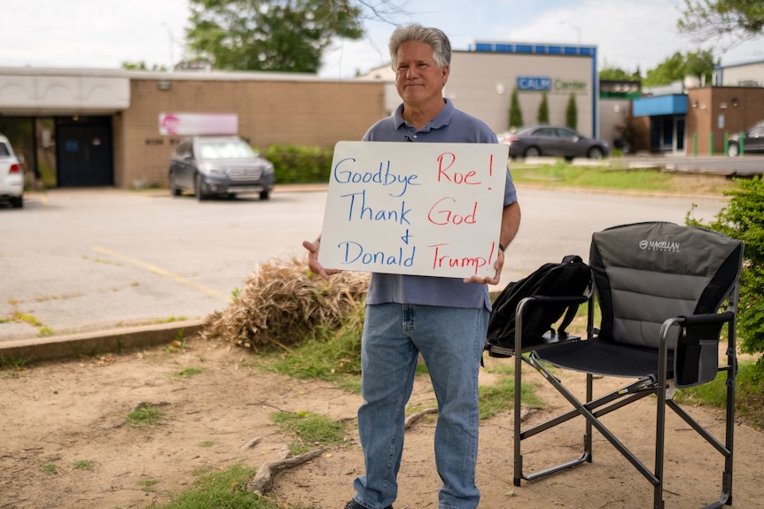 A middle-aged man stands next to a camping chair in a parking lot holding a sign: 'Goodbye Roe Thank God and Donald Trump'