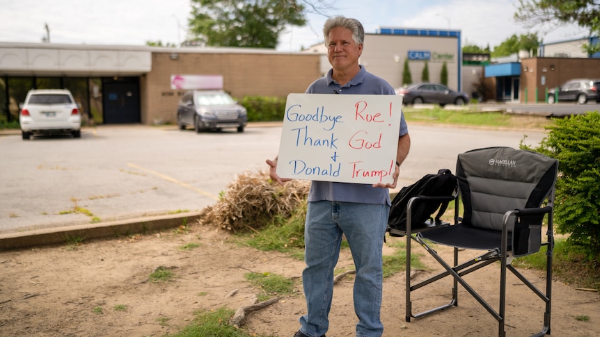 A middle-aged man stands next to a camping chair in a parking lot holding a sign: 'Goodbye Roe Thank God and Donald Trump'