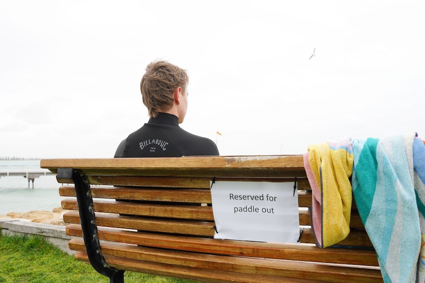 A boy in a wetsuit sits on a bench overlooking the ocean.