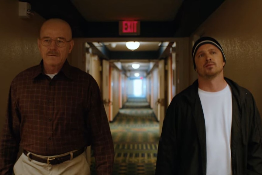 Two men walk down a hotel corridor with an exit sign behind them