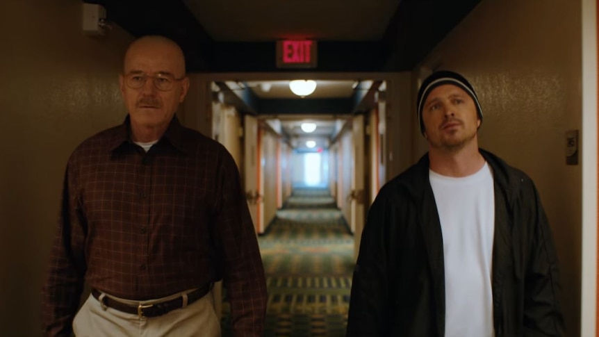 Two men walk down a hotel corridor with an exit sign behind them