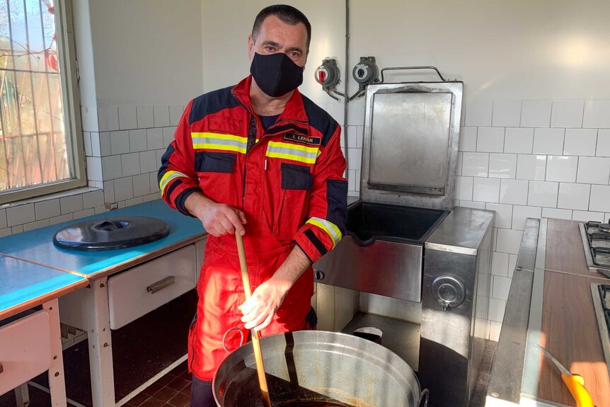 A man in a face mask holding a ladle in a giant pot of goulash.