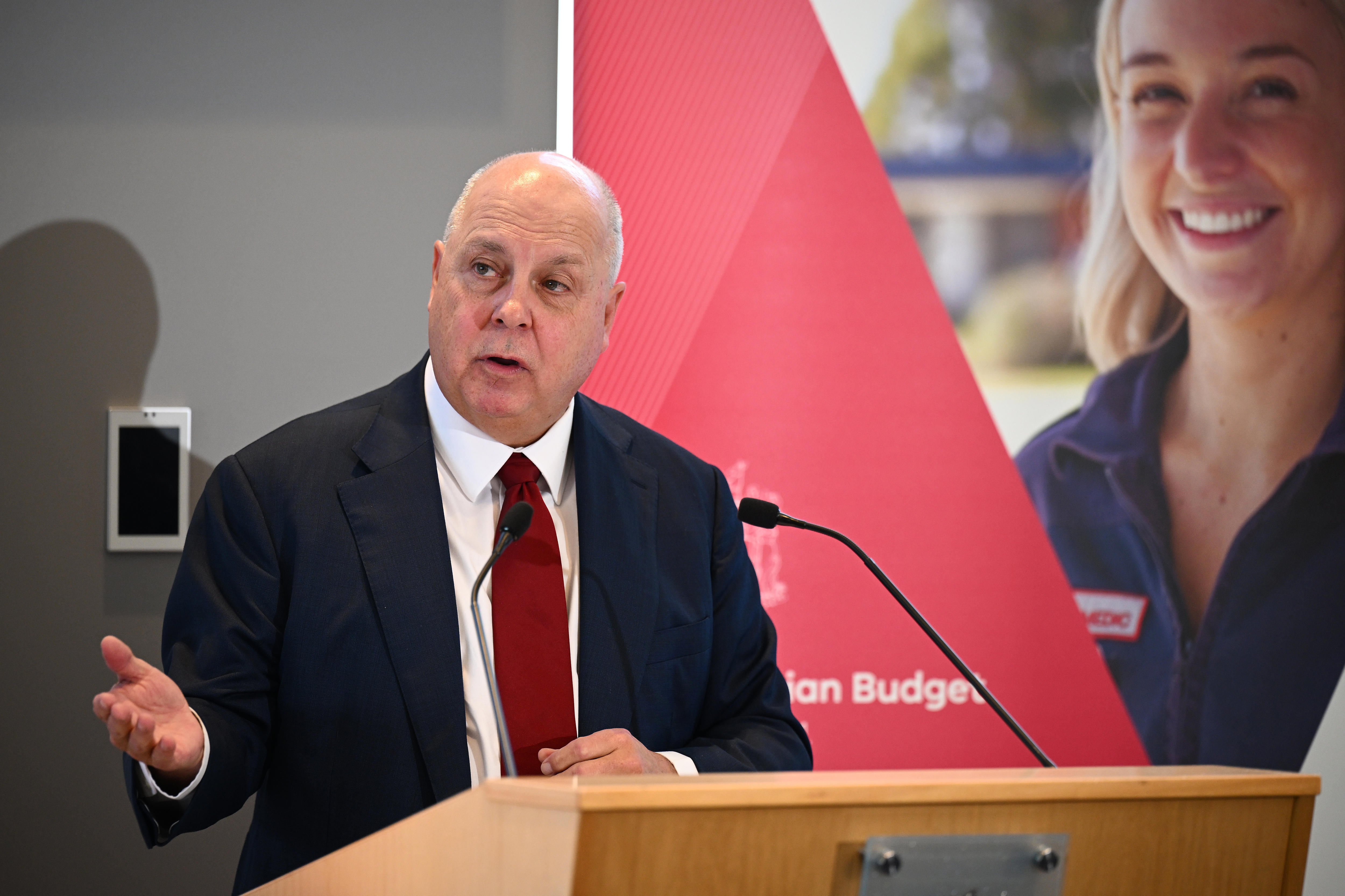 Tim Pallas gestures with his hand as he delivers a speech inside in front of a red backdrop.