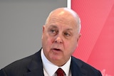 Tim Pallas gestures with his hand as he delivers a speech inside in front of a red backdrop.