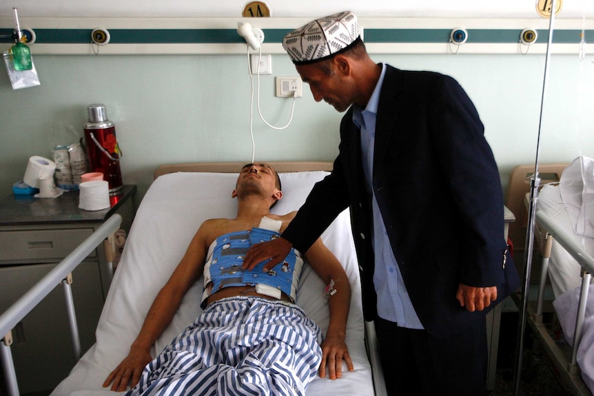 Man wearing traditional Uyghur Muslim hat touches his son who is lying with bandages on his chest in a hospital.
