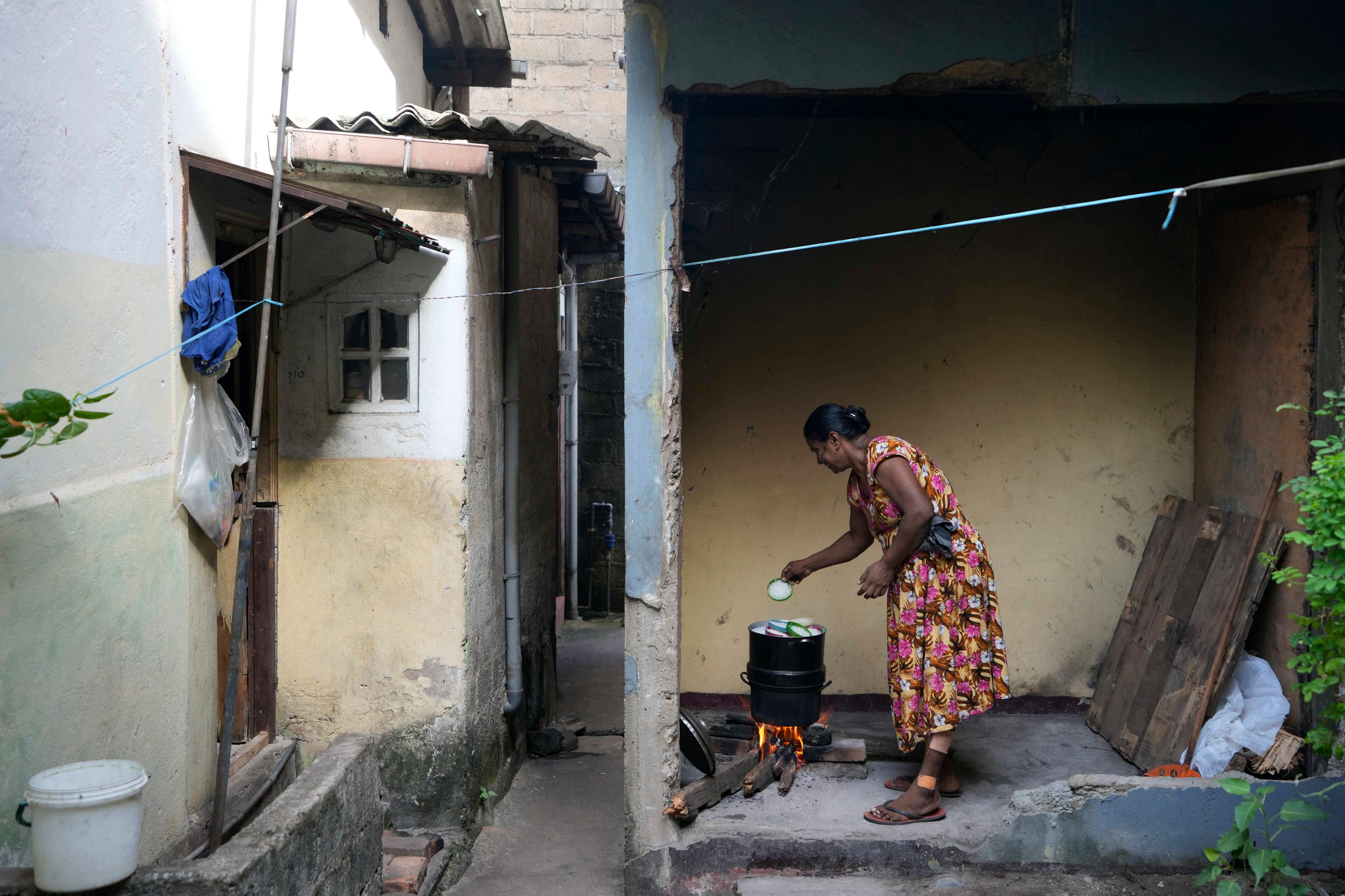 A woman cooks using a firewood hearth outside her house to the right of a small alleyway of houses. 