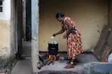 A woman cooks using a firewood hearth outside her house to the right of a small alleyway of houses. 