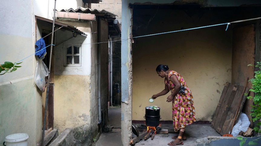 A woman cooks using a firewood hearth outside her house to the right of a small alleyway of houses. 