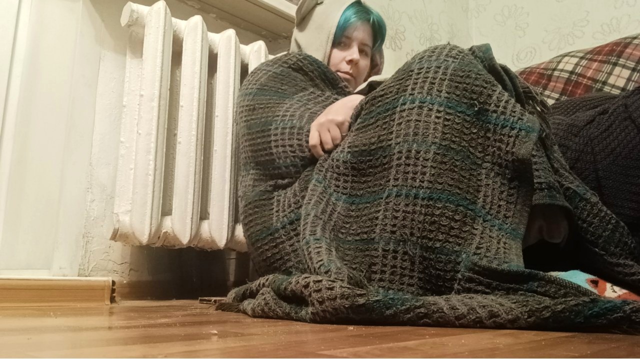A man wrapped in a blanket sits on the floor and frowns at the camera.