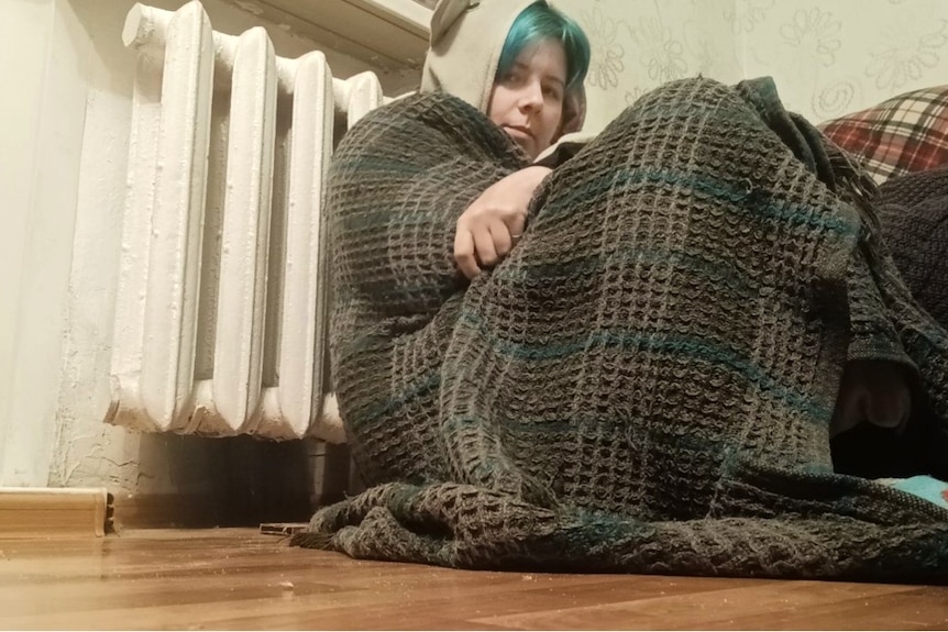 A man wrapped in a blanket sits on the floor and frowns at the camera.