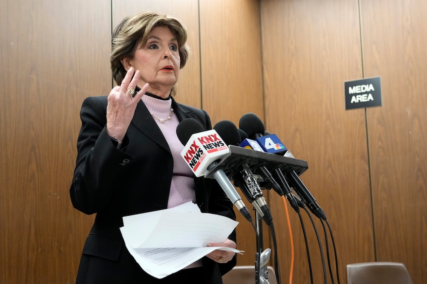 A woman wearing a pink mock turtle neck and black blazer raises three fingers while speaking into several microphones