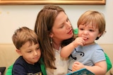 Kate Ellis sits on a couch wearing a green jacket with her two sons on her lap reading a book.