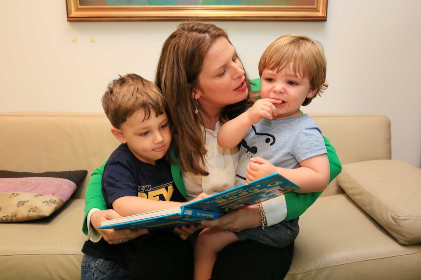 Kate Ellis sits on a couch wearing a green jacket with her two sons on her lap reading a book.