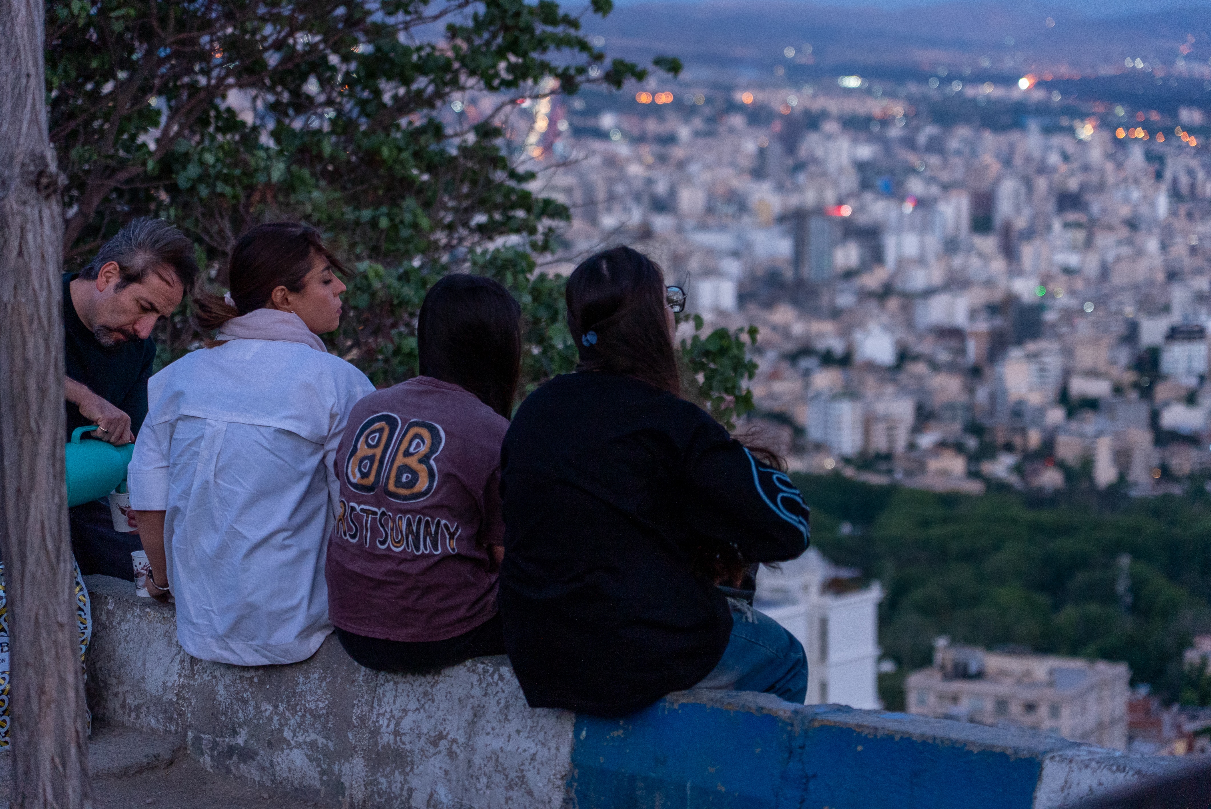 Four people sit at a lookout where the city of Tehran can be seen below them.