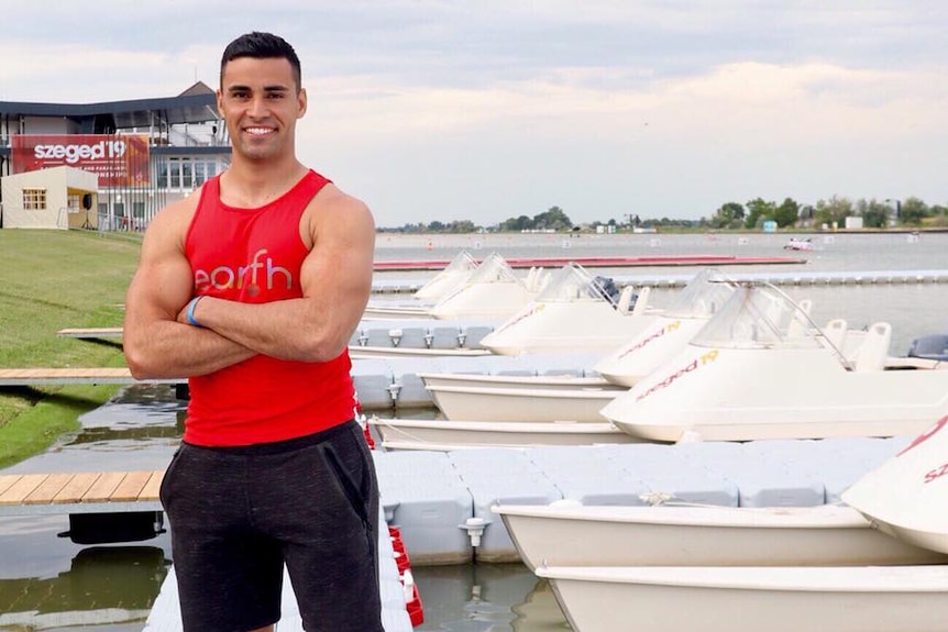 Pita Taufatofua pictured in a red singlet and shorts as he stands on a river jetty with boats moored.